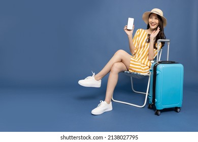 Happy Asian Woman Sitting On Chair With Suitcase And Showing Mobile Phone Application Isolated On Blue Background, Tourist Girl Having Cheerful Holiday Trip Concept