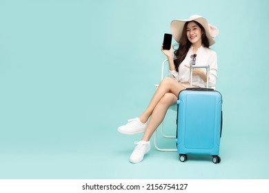 Happy Asian Woman Sitting On Chair With Suitcase And Showing Screen Mobile Phone Isolated On Green Background, Tourist Girl Having Cheerful Holiday Trip Concept