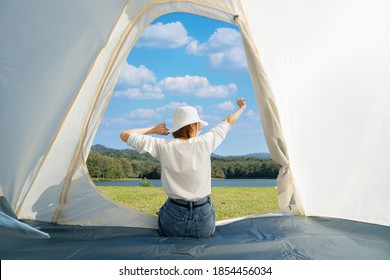 Happy Asian woman sitting with stretching her arms in a camping tent while admiring beautiful calm lake with dense woods and clear blue sky with white clouds in front of her during a warm sunny day. - Powered by Shutterstock