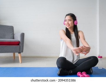 Happy Asian Woman In Sportswear Sitting On Mat Doing Warm Up And Yoga Stretching In Living Room At Home. Young Sports Girl Wears Pink Headphones And Music Listen With Workout Exercise For Healthy Body