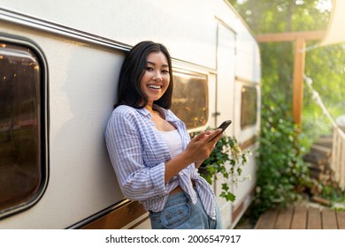 Happy Asian woman standing near camper van, using smartphone on camping trip in countryside, copy space. Cool young lady browsing web, chatting online, watching video on summer vacation outdoors - Powered by Shutterstock