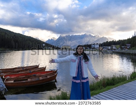 happy Asian woman standing on the coast of Misurina lake. Dolomites, Italy. Landscape with girl, reflection in water, buildings, blue sunset sky. Italian alps. Travel background.