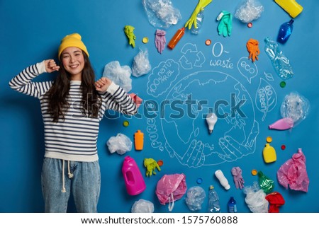Happy Asian woman stretches hands, stands relaxed and satisfied, glad to finish cleaning territory from wastes, poses against blue background, much plastic garbage around. Problem of environment