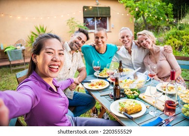 Happy Asian Woman Taking A Selfie Photo Using Smartphone With Diverse Adult Friends Posing For The Picture. A Multiethnic Group Of Men And Women Celebrating A Evening Dinner Party Reunion Together