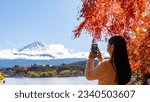 Happy Asian woman travel Japan on autumn holiday vacation. Attractive girl using mobile phone taking selfie during travel the lake around Mt.Fuji and looking beautiful red maple tree leaf falling.