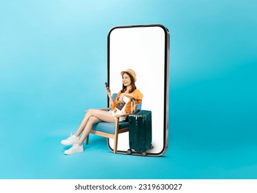 Happy Asian woman traveler sitting on the chair and big smartphone with mockup of blank screen, hand holding passport and plane tickets with suitcase isolated on blue background. - Powered by Shutterstock