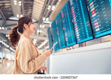 Happy asian woman traveller checking flight schedule departures board in airport terminal hall in front of check in counters. Tourist journey trip concept - Shutterstock ID 2204007367