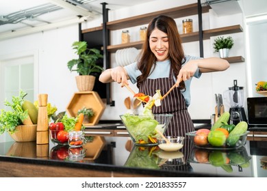 Happy Asian woman wearing apron, tossing the vegetarian salad with wooden spatulas. Preparing a healthy salad with fresh vegetables such as carrot, tomato cabbage and green oak in the home kitchen. - Shutterstock ID 2201833557