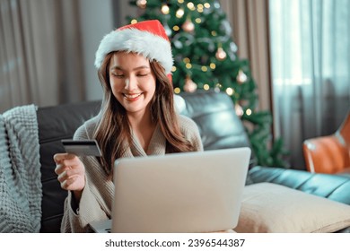 Happy asian woman wearing a Santa hat and holding a phone and a credit card payment shopping Christmas present sitting on couch near decorated Christmas tree, New year celebration. - Shutterstock ID 2396544387