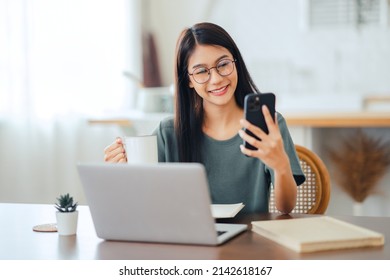 Happy Asian Woman Works At Home And Uses A Smart Phone And A Notebook Laptop Computer On Table. Smiling Young Female Wearing Eyeglasses Messaging With Smartphone.