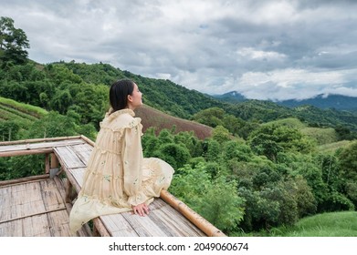 Happy Asian Woman In Yellow Dress Sitting At Wooden Terrace Among The Mountain On Vacation