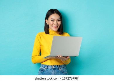 Happy Asian Woman In Yellow Pullover Using Laptop, Shopping Online Or Working, Standing Over Blue Background