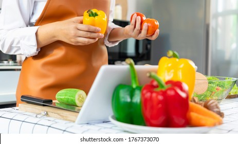 Happy Asian Women Cooking Healthy Food While Looking Recipe On Digital Tablet In The Loft Kitchen Browsing Internet Website On Tutor Cooking Class, Healthy Food And Dieting Concept