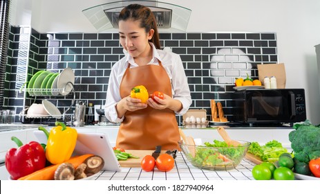 Happy Asian Women Cooking Healthy Food While Looking Recipe On Digital Tablet In The Loft Kitchen Browsing Internet Website On Tutor Cooking Class, Healthy Food And Dieting Concept