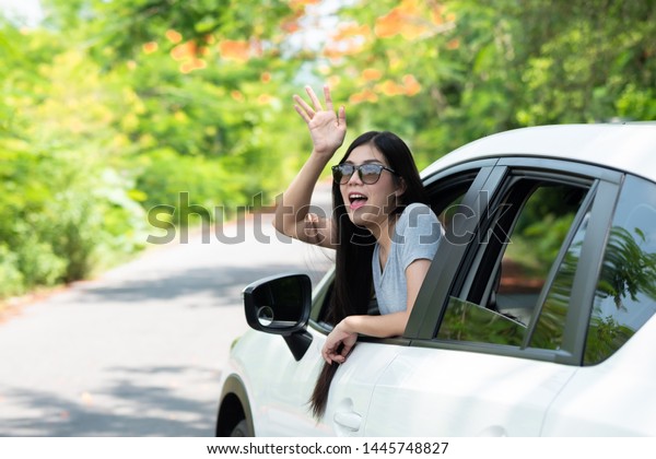 Happy asian women raising hands\
to summer holiday travel trip in the car on the road with sunlight,\
daylight and tree natural background. tourist vacation\
concept