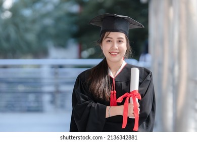 Happy Asian Young Beautiful Graduate Female Student With University Degree Standing And Holding Diploma In Hand After Graduation Wearing Black Cap With Red Tassels. Blur Background Of College Building