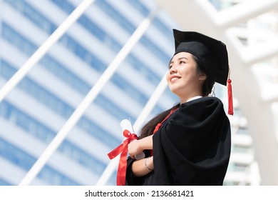 Happy Asian Young Beautiful Graduate Female Student With University Degree Standing And Holding Diploma In Hand After Graduation Wearing Black Cap With Red Tassels. Blur Background Of College Building