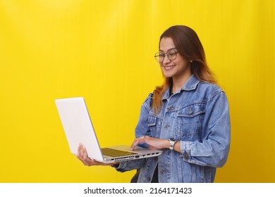 Happy Asian Young College Student Standing While Holding A Laptop. Isolated On Yellow Background