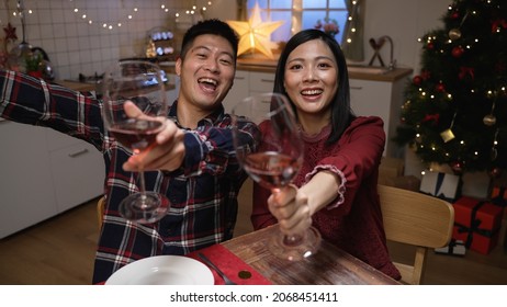 Happy Asian Young Couple Looking At Mobile Cam And Making Christmas Toast To Friend With Red Wine During A Virtual Chat In A Cozy Home At Dinner.