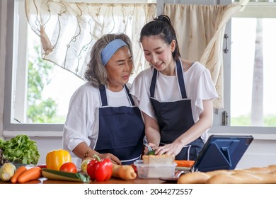 Happy Asian Young Daughter And Senior Mother Cooking Online  Class On Tablet Together Making Fresh Vegetables Food In Kitchen At Home