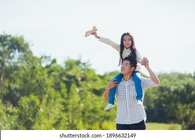 Happy Asian Young Family Father And Carrying An Excited Girl On Shoulders Having Fun And Enjoying Outdoor Lifestyle Together Playing Aircraft Toy On Sunny Summer Day, Father's Day Concept