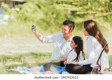 Happy Asian Young Family Father, Mother And Child Little Girl Having Fun And Enjoying Outdoor Sitting On Picnic Blanket Taking Selfie Using Technology Mobile Smart Phone At Summer Garden Park