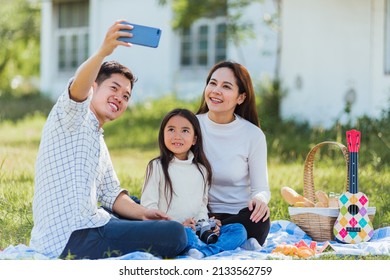 Happy Asian Young Family Father, Mother And Child Little Girl Having Fun And Enjoying Outdoor Sitting On Picnic Blanket Taking Selfie Using Technology Mobile Smart Phone At Summer Garden Park