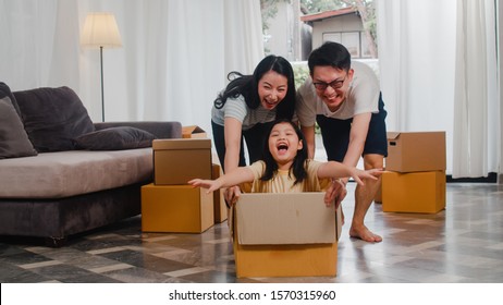 Happy Asian Young Family Having Fun Laughing Moving Into New Home. Japanese Parents Mother And Father Smiling Helping Excited Little Girl Riding Sitting In Cardboard Box. New Property And Relocation.