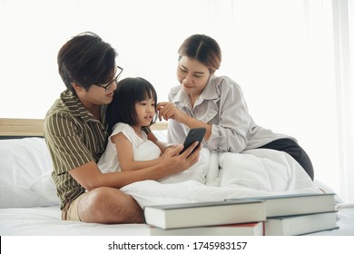 Happy Asian Young Family Spend Time Together. Dad Hold Mobile Phone Taking Photo Selfie With Mom Daughter Who Sitting Under White Blanket On The Bed At Bedroom