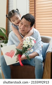 Happy Asian Young Father Getting Bunch Of Flowers And Handmade Card Congratulations On Fathers Day From Little Cute Daughter. Dad Is Piggyback Ride Kid With Love In Living Room At Home