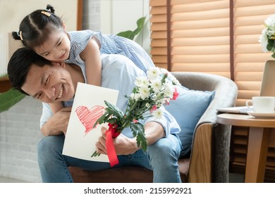 Happy Asian Young Father Getting Bunch Of Flowers And Handmade Card Congratulations On Fathers Day From Little Cute Daughter. Dad Is Piggyback Ride Kid With Love In Living Room At Home