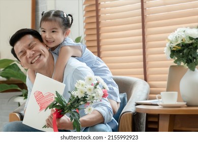 Happy Asian Young Father Getting Bunch Of Flowers And Handmade Card Congratulations On Fathers Day From Little Cute Daughter. Dad Is Piggyback Ride Kid With Love In Living Room At Home