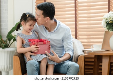 Happy Asian Young Father Kissing Forehead Of His Little Cute Daughter Who Surprise Give Him Red Gift Box And Sitting On Lap While He Is Working With Laptop In Living Room At Home On Father's Day