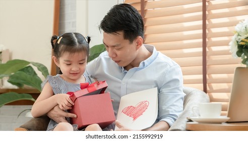 Happy Asian Young Father Surprise With Red Gift Box And Heart Colored Card Which Little Cute Daughter Give Him And Sitting On Lap While He Is Working With Laptop In Living Room At Home On Father's Day