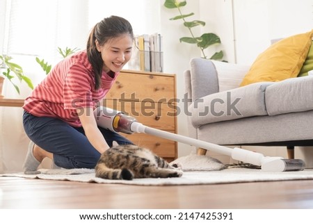 Happy asian young housekeeper woman using vacuum cleaning, cleaner to remove dust, hair or fur on floor in living room while cute cat lying on carpet. Routine housework, chore in household of maid.