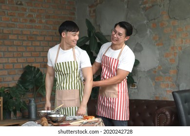 Happy Asian Young LGBT Gay Couple Making Pizza Together. A Man Helping His Boyfriend Put On An Apron In Kitchen At Home. Relationship Of Homosexual Lifestyle Concept