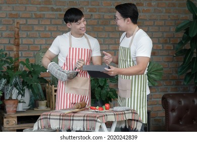 Happy Asian Young LGBT Gay Couple With Apron Cooking Together. Handsome Man Mixing Salad Dressing And His Boyfriend Thumb Up To Him And Using Tablet In Kitchen At Home.