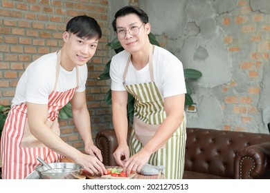 Happy Asian Young LGBT Gay Couple With Apron Cooking Together. Handsome Men Cutting Tomato Cucumber To Make Salad In Kitchen At Home. Relationship Of Homosexual Lifestyle Concept