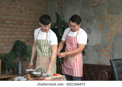 Happy Asian Young LGBT Gay Couple Making Pizza Together. A Man Helping His Boyfriend Put On An Apron In Kitchen At Home. Relationship Of Homosexual Lifestyle Concept