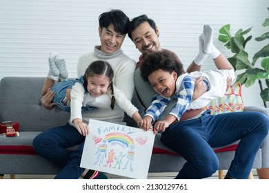 Happy Asian young LGBTQ gay couple playing carrying little cute adopted Caucasian   African kid in hands   showing rainbow family drawing in living room at home  LGBT diverse family concept