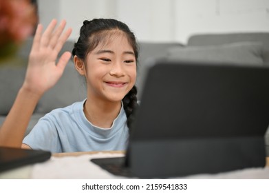 Happy Asian Young Little Girl Saying Hi Greeting Her Friends And Teacher, Enjoy Studying On Online Class Via Tablet At Home.