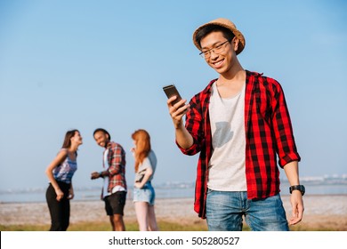 Happy Asian Young Man Standing And Using Mobile Phone While His Friends Talking Outdoors