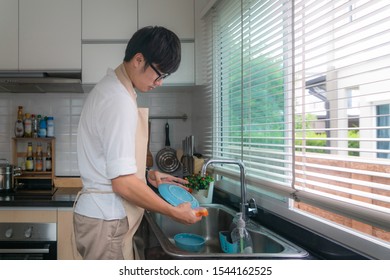 Happy Asian Young Man In White Shirt Standing And Washing Dishes In Sink On The Kitchen. Kitchen Ornaments And Kitchenware Supplies In Background. Man Doing Housework In House Concept.