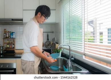 Happy Asian Young Man In White Shirt Standing And Washing Dishes In Sink On The Kitchen. Kitchen Ornaments And Kitchenware Supplies In Background. Man Doing Housework In House Concept.