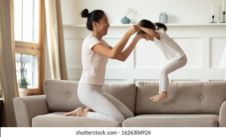 Happy Asian Young Mother Rest On Sofa In Living Room Play With Little Biracial Daughter, Overjoyed Vietnamese Mom Or Nanny Have Fun Jump Engaged In Funny Game With Small Ethnic Girl Child At Home