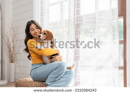 Happy Asian young woman in casual clothes hugging her beagle dog sitting near window in living room at cozy home. Pet and cute animal concept.