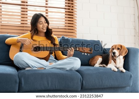 Happy Asian young woman in casual clothes singing and playing guitar with beagle dog on sofa in living room at home