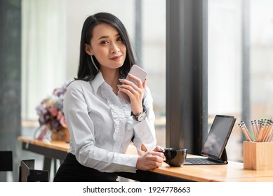 Happy Asian Young Woman Drinking Coffee At Coffee Shop Holding Phone. Looking At Camera.