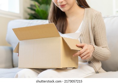 Happy asian young woman, girl customer sitting on sofa at home, hand in opening and unpacking cardboard box carton parcel after buying ordering present, shopping online, delivery service concept.