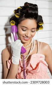 happy and asian young woman with hair curlers standing in pink ruffled top, pearl necklace and white gloves and talking on purple retro phone and smiling near white tiles, housewife
